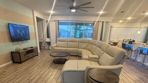 A modern living room with a sectional sofa, a flat-screen TV, wooden flooring, and a ceiling fan.