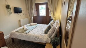 Un dormitorio con una cama, una mesita de noche con una planta, un espejo, un televisor y una ventana con cortinas.