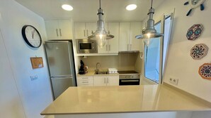 A modern kitchen with white cabinetry, a stainless steel refrigerator, a microwave, and a built-in oven. There are pendant lights above the island and a clock on the wall. - SUITE CON VISTAS A PLAYA DE SON BOU (Son Bou (Platja))
