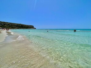 Une plage avec une eau turquoise et limpide, un rivage sablonneux et un promontoire rocheux en arrière-plan.