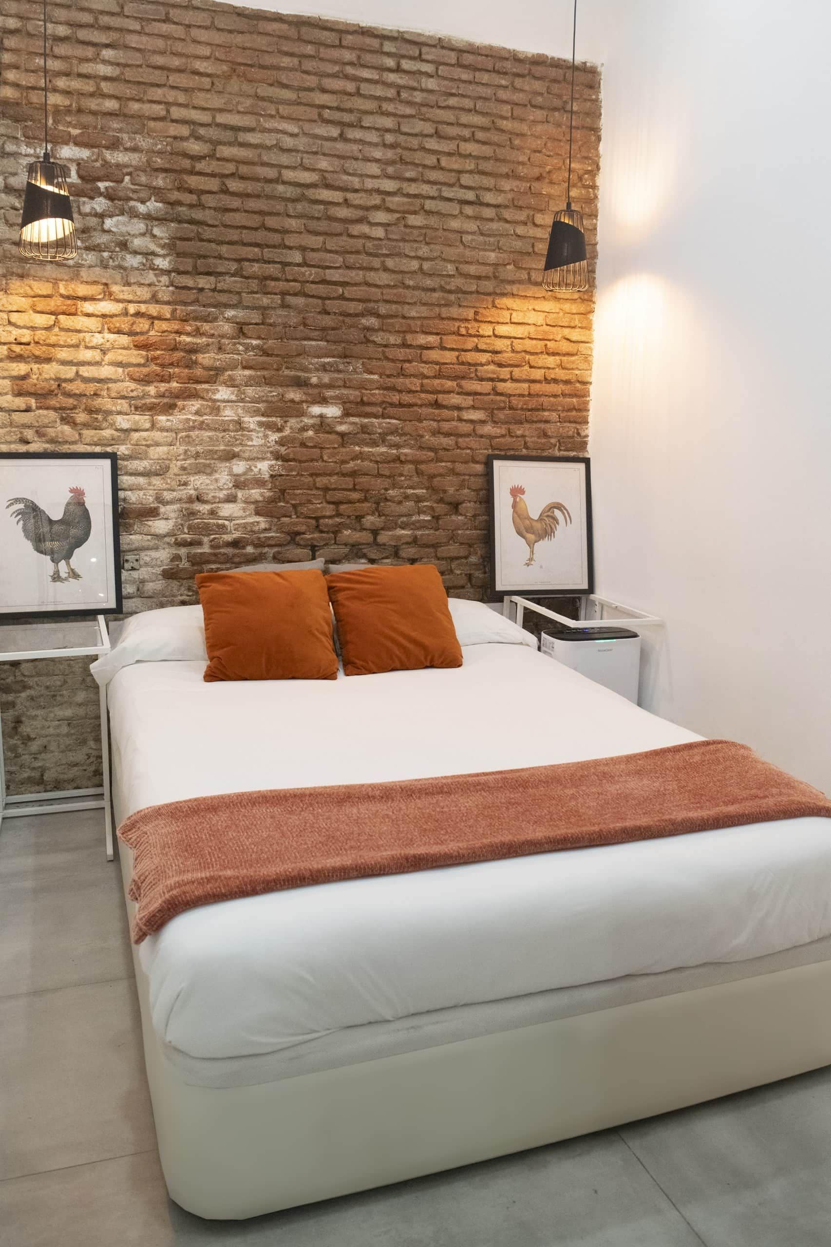 A bedroom with a brick wall, two framed rooster pictures, a bed with white bedding and orange pillows, and two bedside lamps.