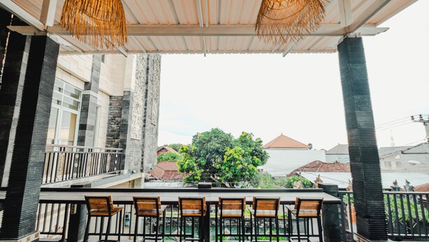A covered outdoor seating area with a bar, wooden chairs, and a view of a tree and buildings in the background. - Hotel Mega Dana (Denpasar)