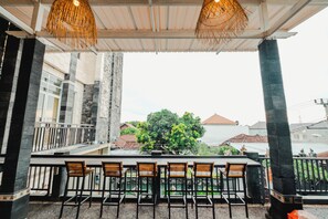 A covered outdoor seating area with a bar, wooden chairs, and a view of a tree and buildings in the background. - Hotel Mega Dana (Denpasar)