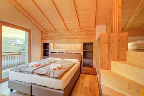 A bedroom with a large bed, wooden ceiling beams, a window with a view of the outdoors, and a built-in bench.