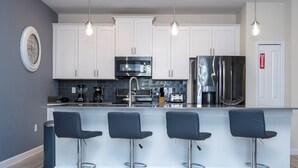 A modern kitchen with white cabinets, stainless steel appliances, and a bar area with four stools. - 4337 QS - Charming Hideaway (Kissimmee)
