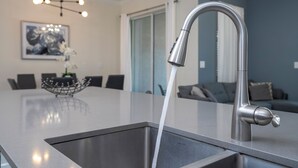 A modern kitchen with a stainless steel faucet, a large island, and a dining area in the background. - 4337 QS - Charming Hideaway (Kissimmee)
