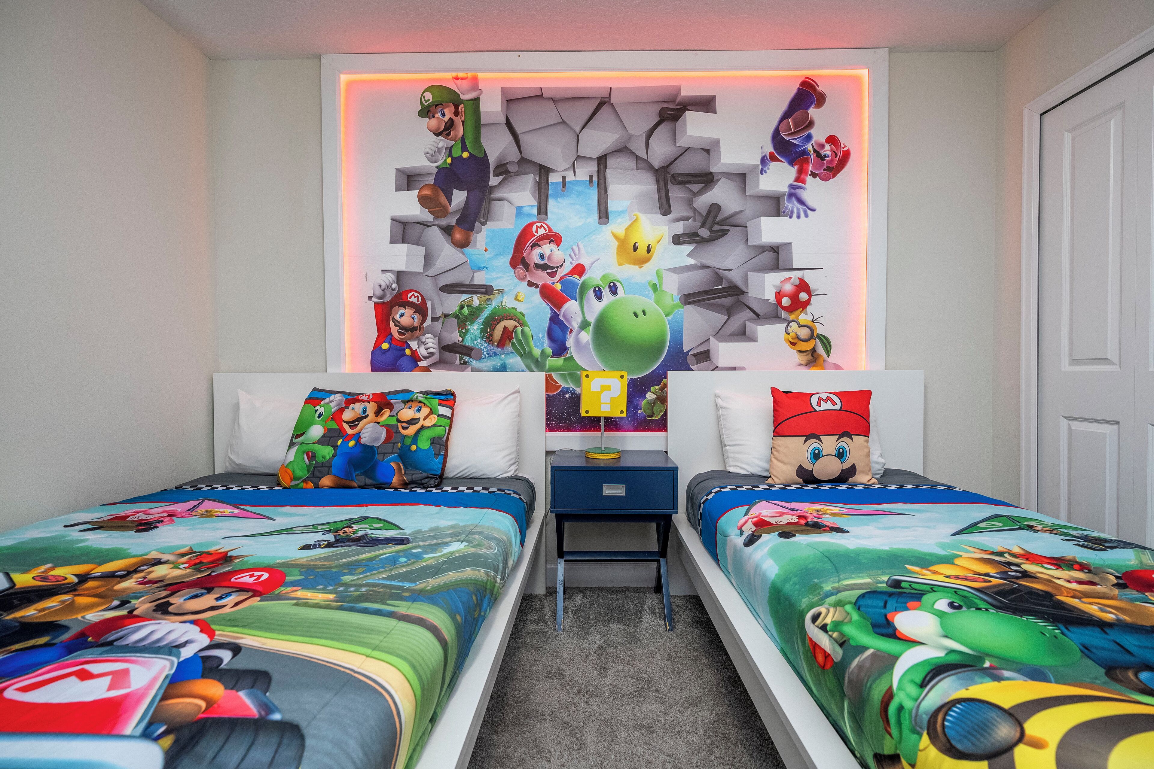 A bedroom with two beds, each adorned with Mario-themed bedding. A wall-mounted shelf displays various Mario and Luigi figurines.