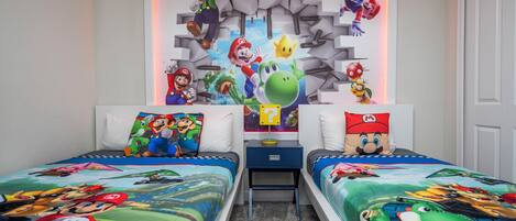 A bedroom with two beds, each adorned with Mario-themed bedding. A wall-mounted shelf displays various Mario and Luigi figurines.