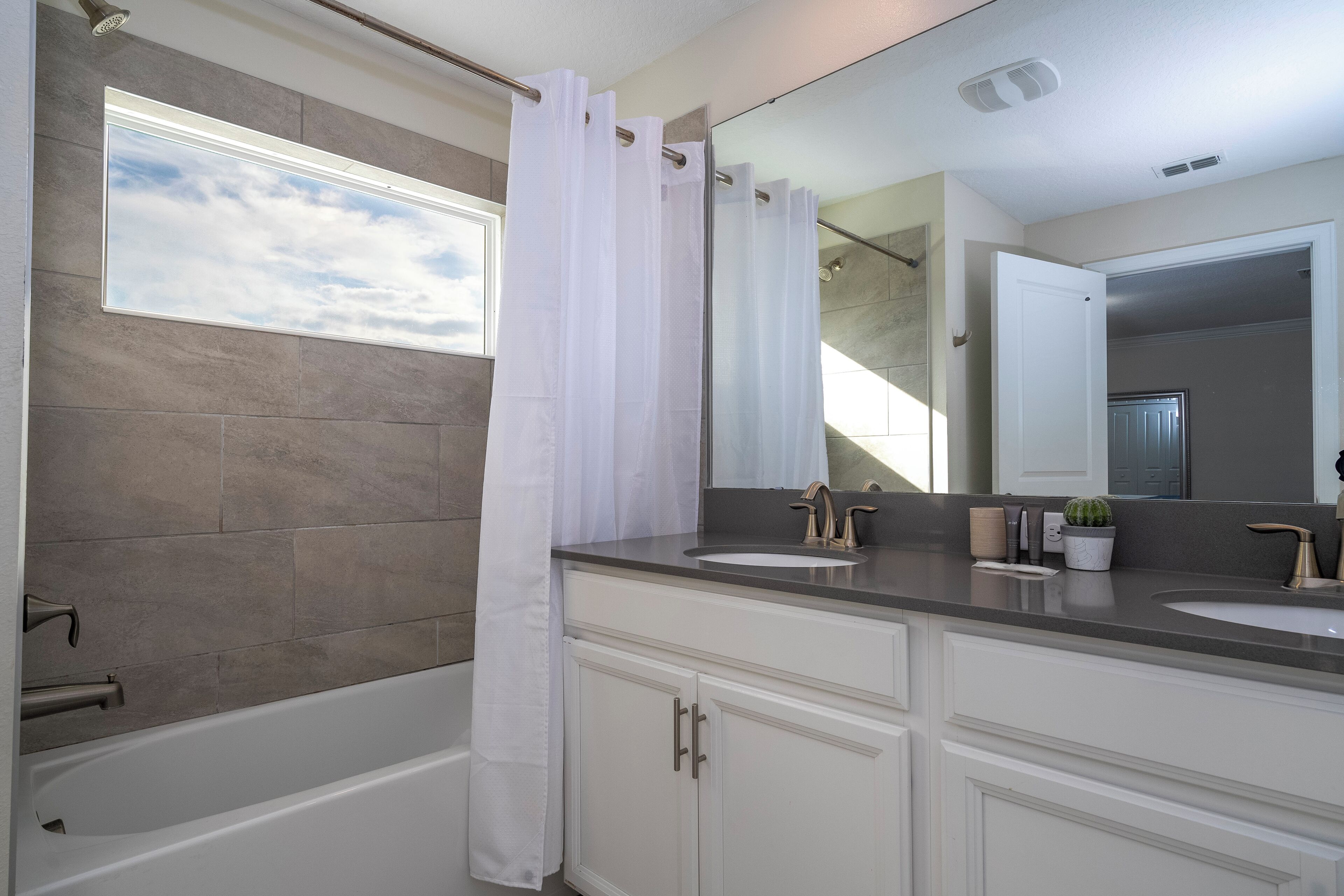 A modern bathroom with a bathtub, two sinks, a large mirror, and a window with a view of the sky.