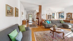 Ein geräumiges Wohnzimmer mit grauem Sofa, einem großen Ecksofa und einem Couchtisch. An den Wänden hängen Bilder in Rahmen, und im Hintergrund ist eine Küchenzeile zu sehen.