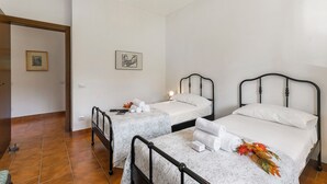 Ein Zimmer mit zwei Betten, beide mit Metallgestell und weißer Bettwäsche. An der Wand hängt ein gerahmtes Bild, und eine Tür führt in einen weiteren Raum.