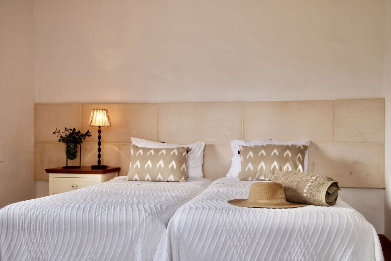 Una cama doble con ropa de cama blanca, dos almohadas estampadas, una lámpara de noche y una planta pequeña en una mesita de noche.
