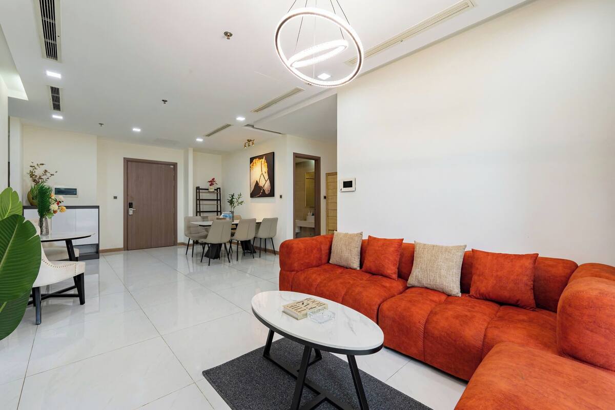 Uma sala de estar moderna com um sofá seccional, uma área de jantar e uma cozinha ao fundo.