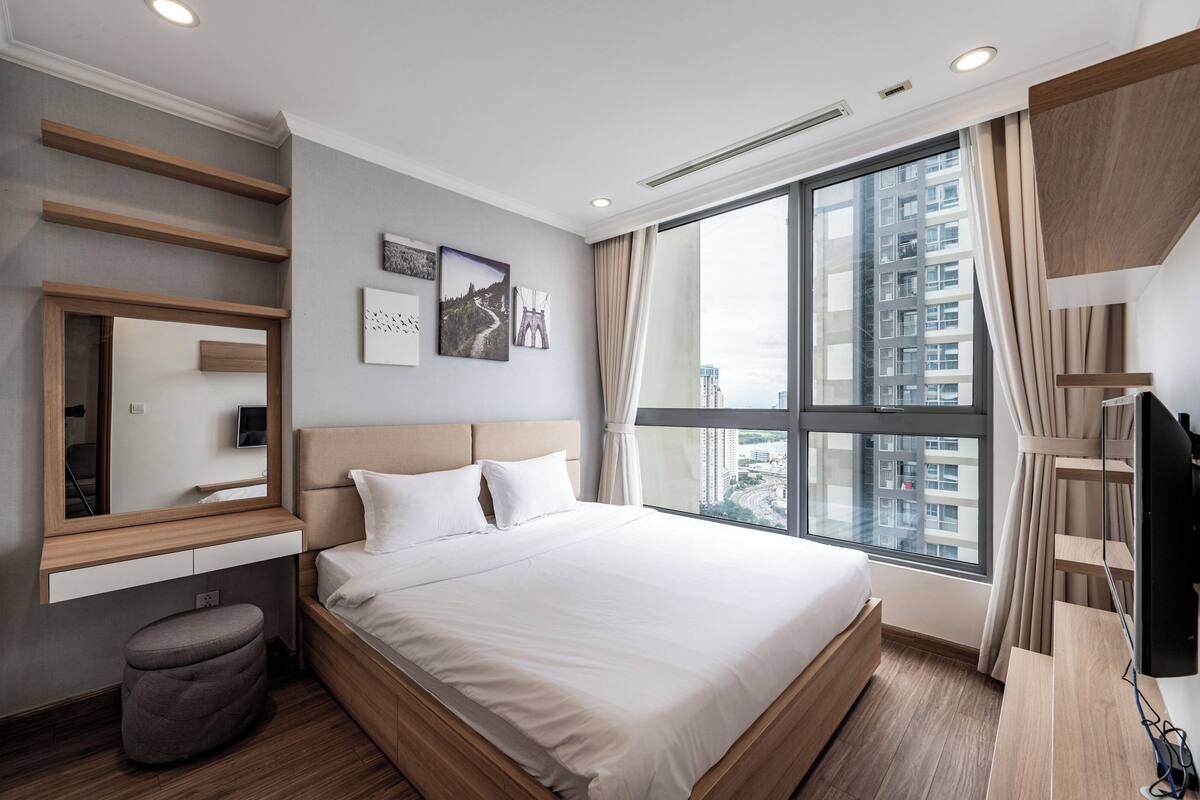 Quarto de hotel moderno com uma cama grande, uma mesa embutida, um banquinho pequeno e uma grande janela com vista para a cidade.