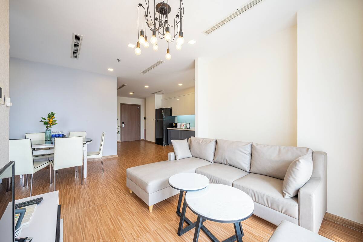 Uma sala de estar moderna com um sofá seccional, uma mesa de jantar com cadeiras e uma cozinha ao fundo.