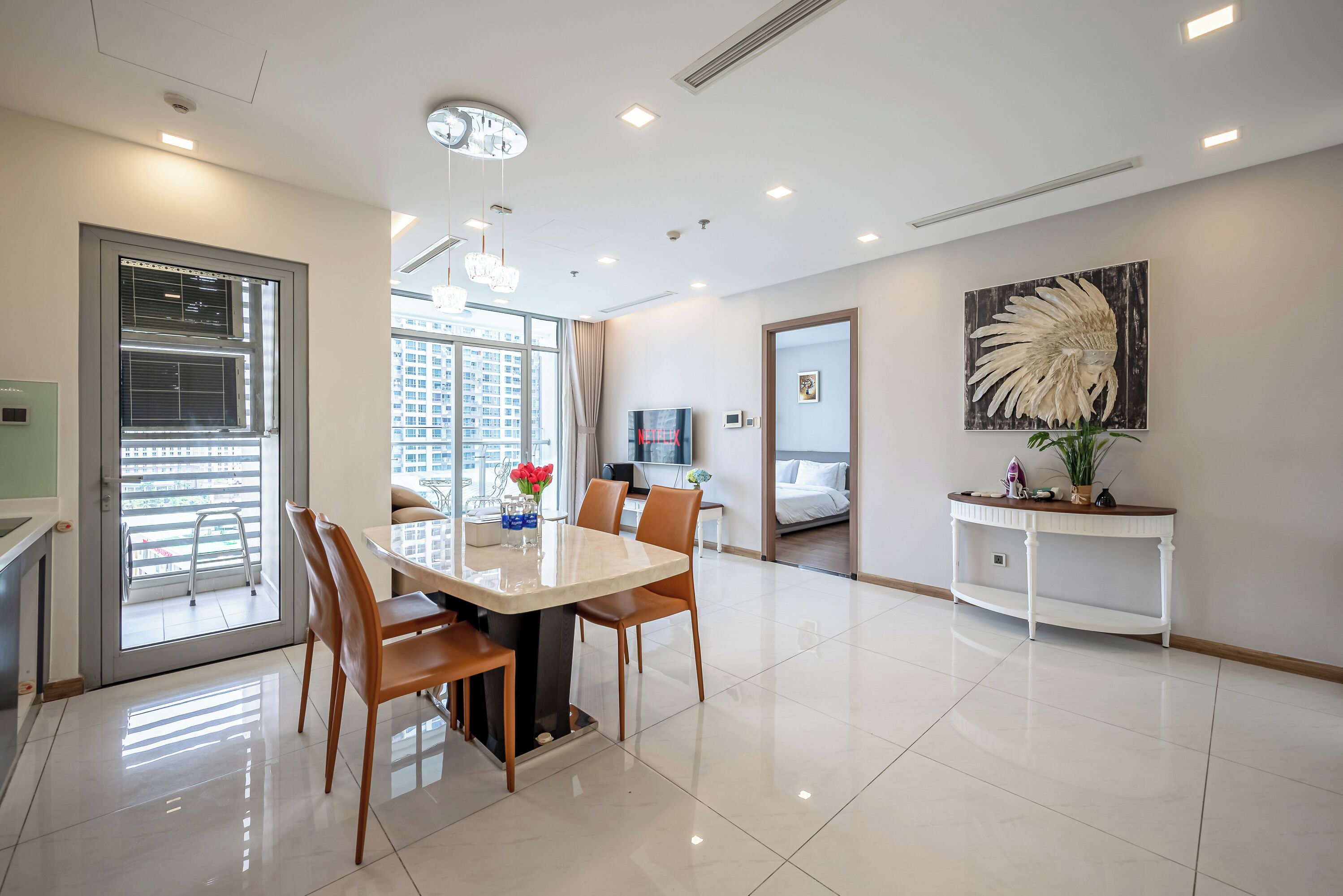 A modern hotel room with a dining area, a large window with a view of the city, a ceiling fan, and a decorative plant on a console table.