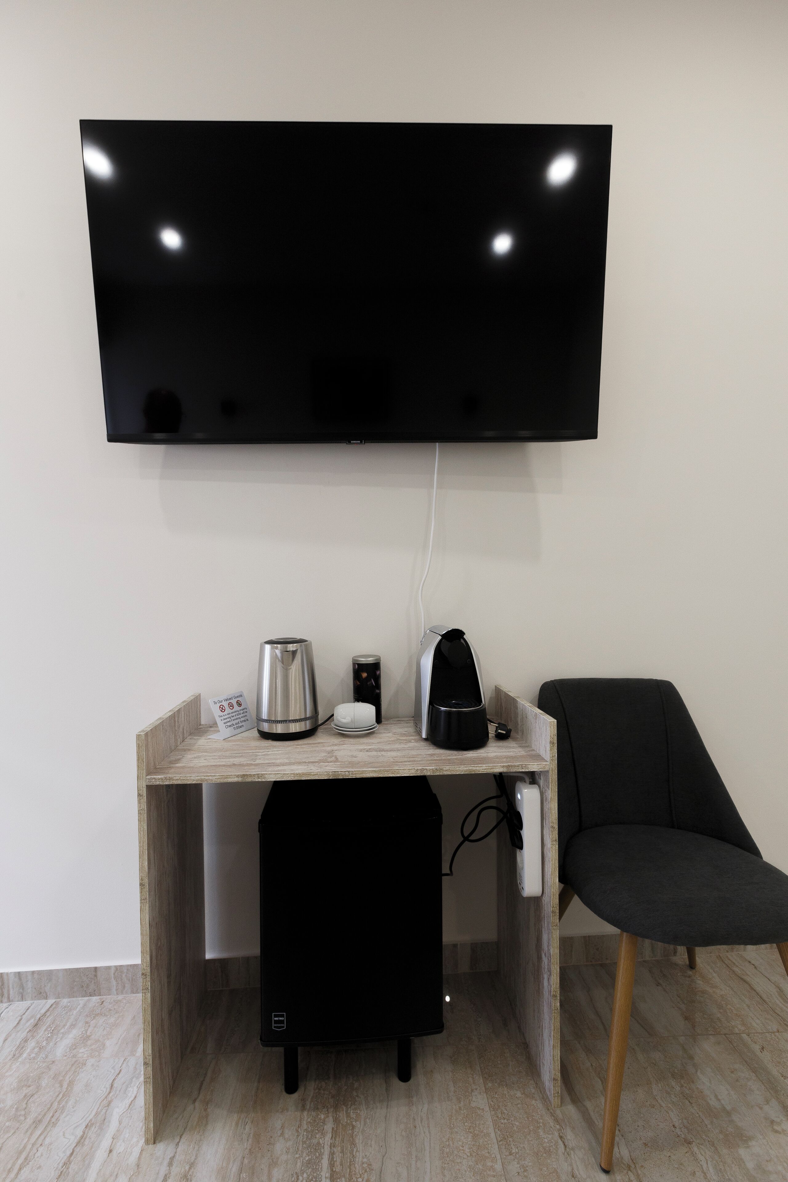A modern room with a flat-screen TV mounted on the wall, a wooden desk with a kettle, a coffee maker, and a black chair.