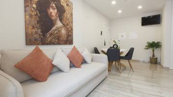Uma sala de estar moderna com um sofá, uma mesa de jantar com cadeiras, uma TV na parede e um vaso com planta.