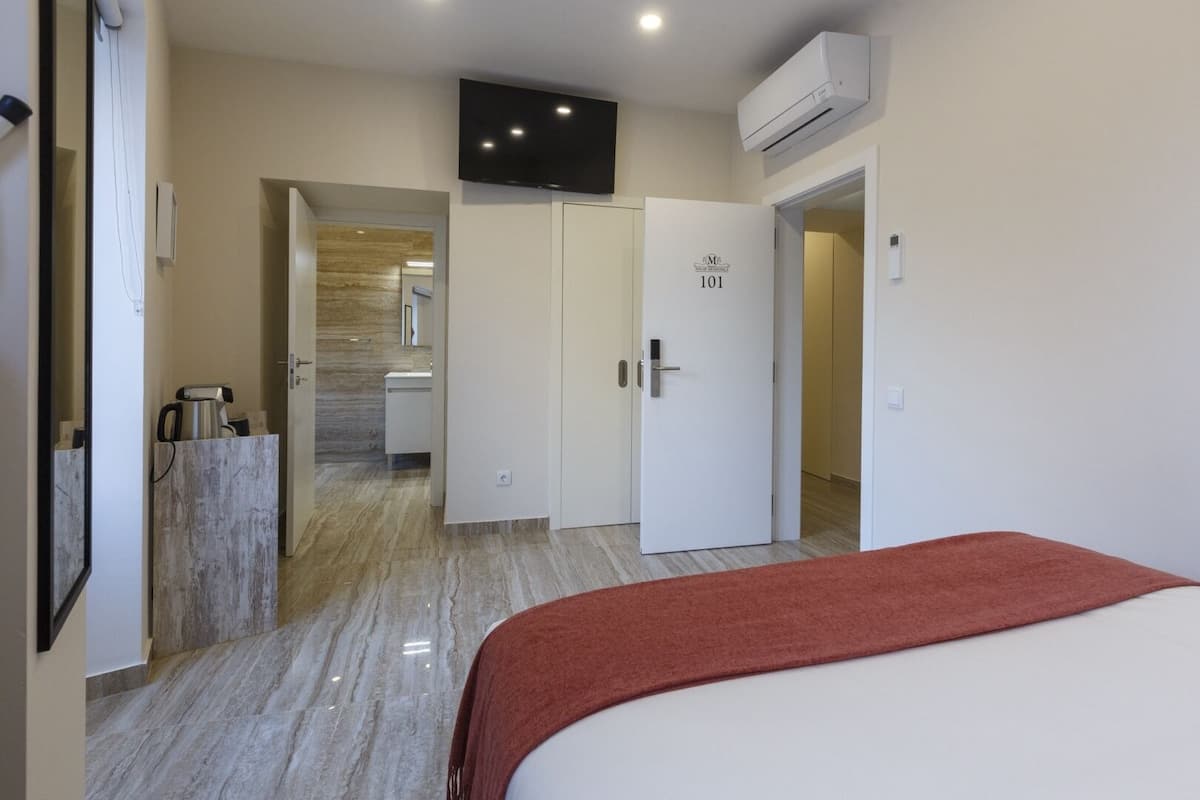 A hotel room with a bed, a TV mounted on the wall, an air conditioning unit, and a bathroom visible through an open door.