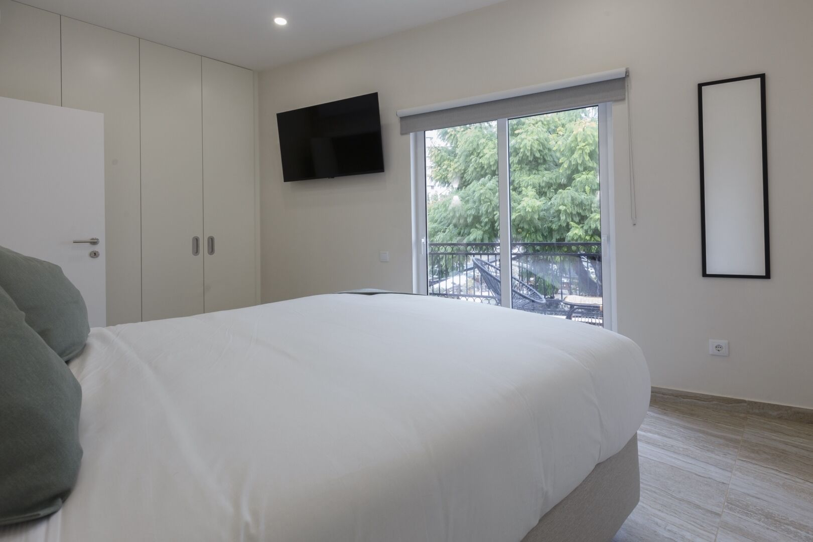 A bedroom with a bed, a TV mounted on the wall, a window with a view of trees, and a door.