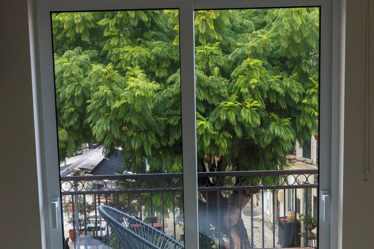 A balcony with a metal chair and a table, a tree in the background, and a building across the street.