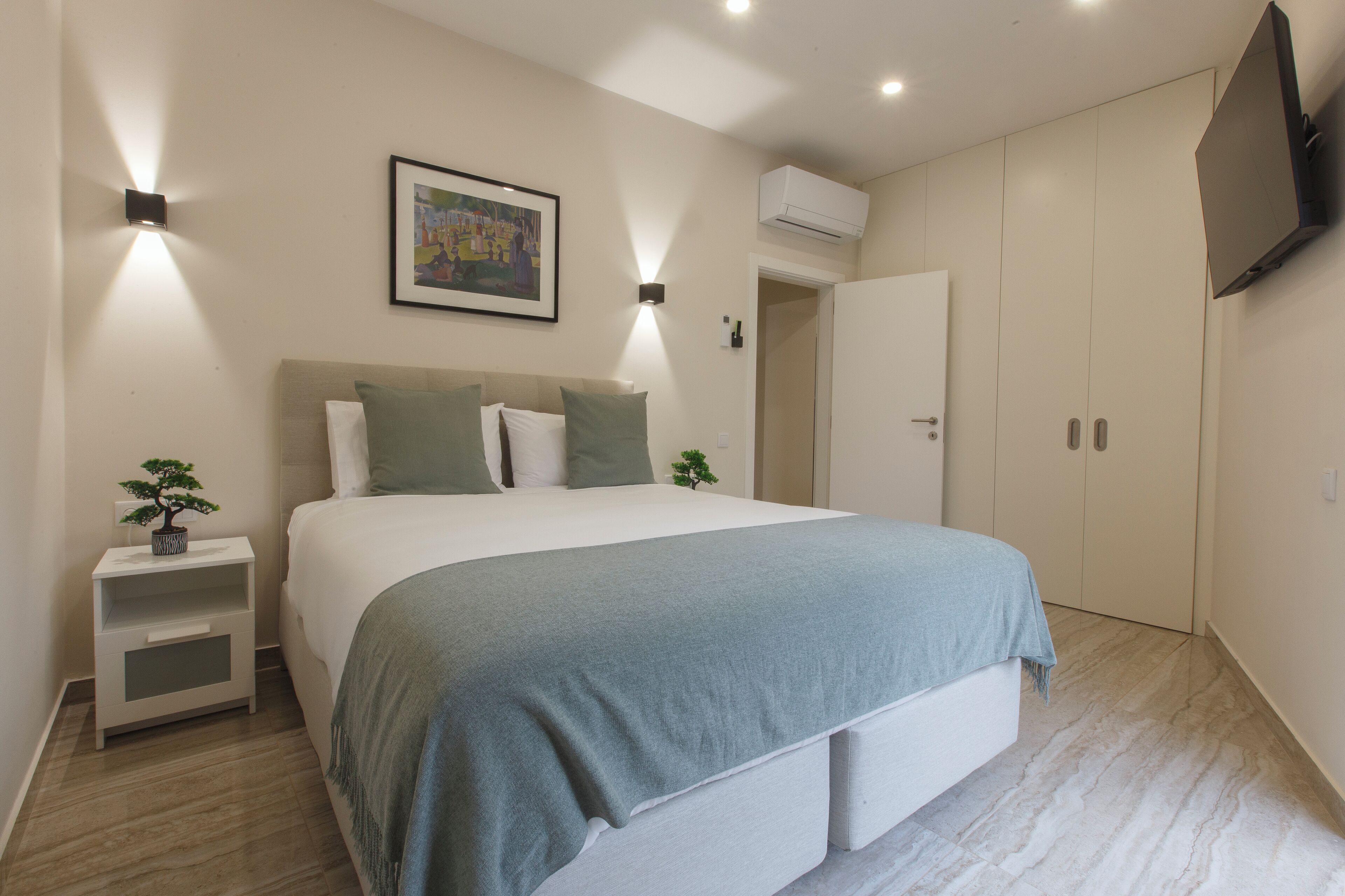Une chambre à coucher comprenant un lit, des tables de chevet, un téléviseur fixé au mur et une porte donnant sur une autre pièce.