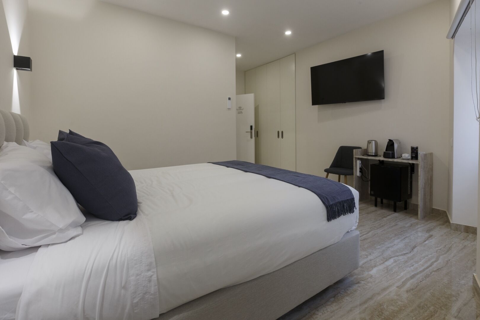 A modern hotel room with a large bed, a flat-screen TV mounted on the wall, a small desk with a coffee maker, and a wardrobe.