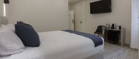 A modern hotel room with a large bed, a flat-screen TV mounted on the wall, a small desk with a coffee maker, and a wardrobe.