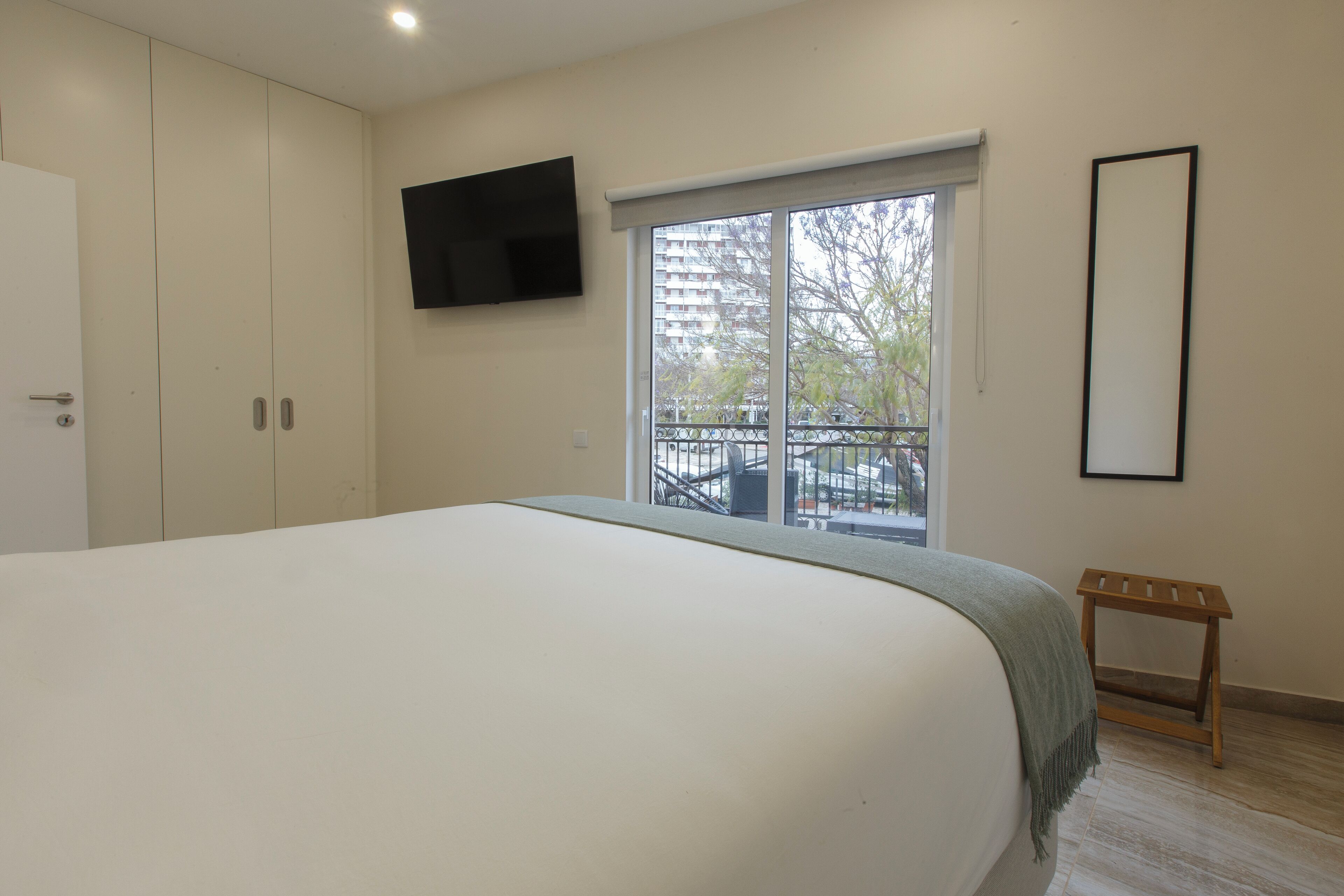 Un dormitorio con una cama grande, un televisor instalado en la pared, una ventana con vista a un balcón y un taburete de madera pequeño.