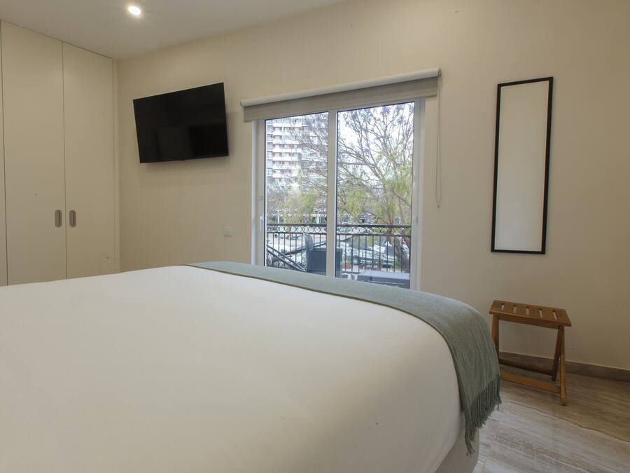 Un dormitorio con una cama grande, un televisor instalado en la pared, una ventana con vista a un balcón y un taburete de madera pequeño.