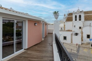 Ein Balkon mit Holzboden, einer Glastür und Blick auf ein weißes Gebäude mit einem Glockenturm.