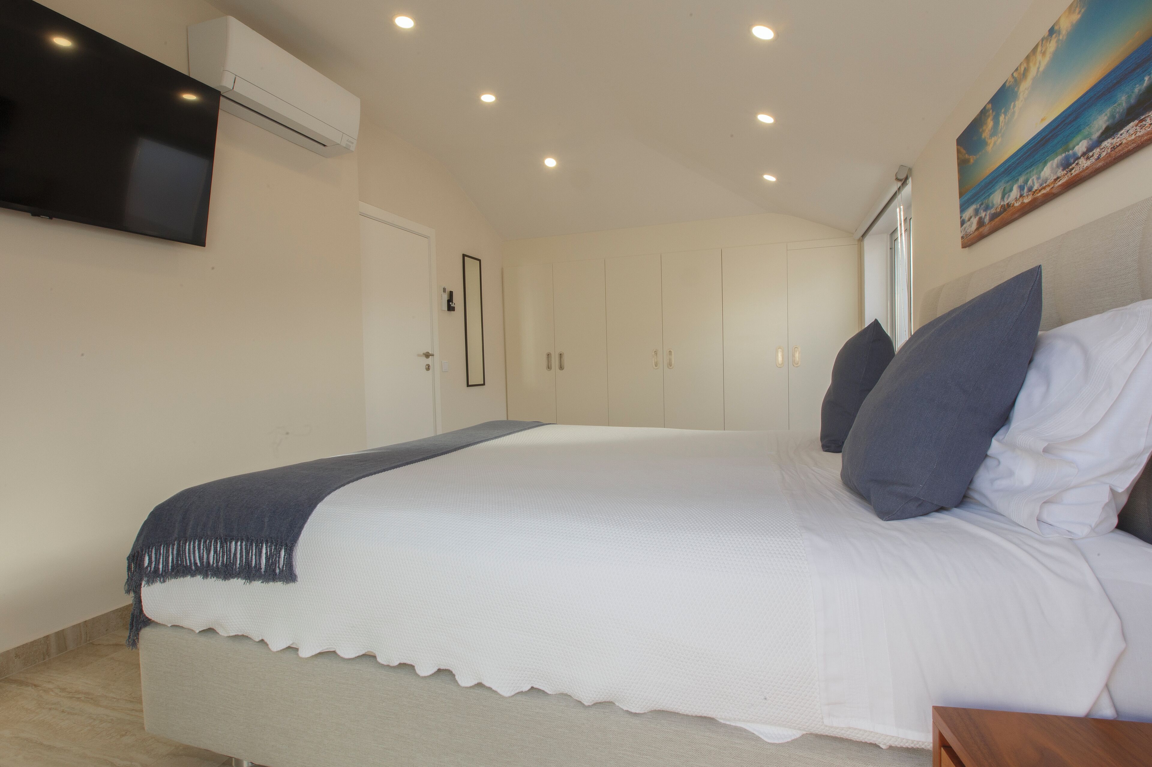 A bedroom with a large bed, a flat-screen TV mounted on the wall, and a painting above the bed.