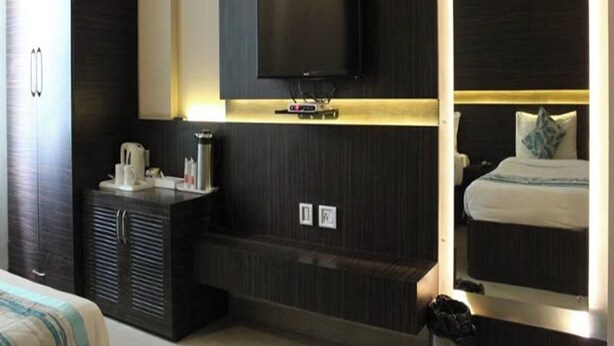 A modern hotel room with a flat-screen TV mounted on a dark wall, a bedside table with a lamp, and a large mirror reflecting the room. - Hotel Ajay International (Prayagraj)