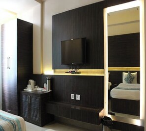 Ein modernes Hotelzimmer mit einem am dunklen Wand montierten Flachbildfernseher, einem Nachttisch mit Lampe und einem großen Spiegel, der den Raum reflektiert.