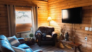 Una habitación acogedora tipo cabaña con un sofá de cuero, un sillón de cuero, un televisor de pantalla plana montado en la pared, una lámpara y una mesita con adornos.