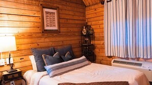 Una habitación de cabaña de madera con una cama, un gráfico enmarcado de peces, una mesita de noche, una lámpara y una ventana con cortinas.