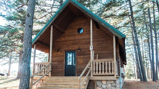 Una cabaña de madera con base de piedra, una puerta principal y un pequeño porche con barandilla.