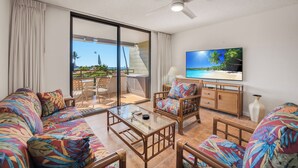 A spacious living room with a sofa, coffee table, and chairs, a television, and a view of the outdoors. - Central AC Ocean & Mountain View 2-bedroom 2-Bath condo across Magic Sands Beach (Kailua-Kona)