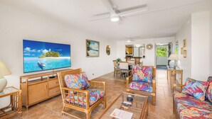 A spacious living area with a television displaying a beach scene, wicker furniture, and a dining area in the background. - Central AC Ocean & Mountain View 2-bedroom 2-Bath condo across Magic Sands Beach (Kailua-Kona)