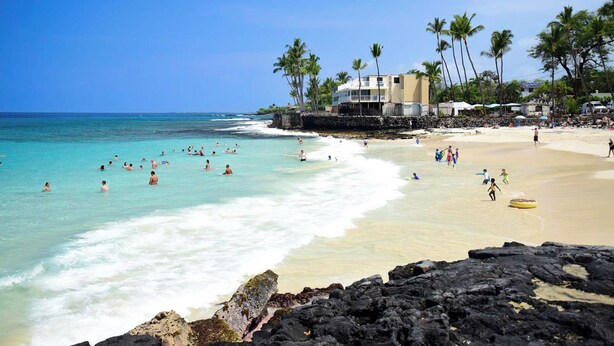 A beach with clear turquoise water, people swimming and sunbathing, and a building with a balcony overlooking the beach. - Central AC Ocean & Mountain View 2-bedroom 2-Bath condo across Magic Sands Beach (Kailua-Kona)