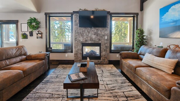 A living room with a stone fireplace, leather sofas, and large windows. - Lucky 7 Lodge – Spacious 4-Bedroom Cabin with Double Decks, Hot Tub & Mountain Views (Lead)