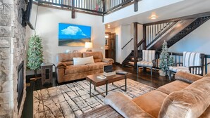 A spacious living area with a fireplace, a large painting, and a staircase leading to an upper level. - Lucky 7 Lodge – Spacious 4-Bedroom Cabin with Double Decks, Hot Tub & Mountain Views (Lead)