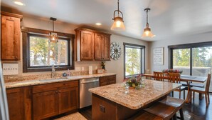 A kitchen with wooden cabinets, granite countertops, a clock, and a dining area with wooden chairs and a table. - Lucky 7 Lodge – Spacious 4-Bedroom Cabin with Double Decks, Hot Tub & Mountain Views (Lead)
