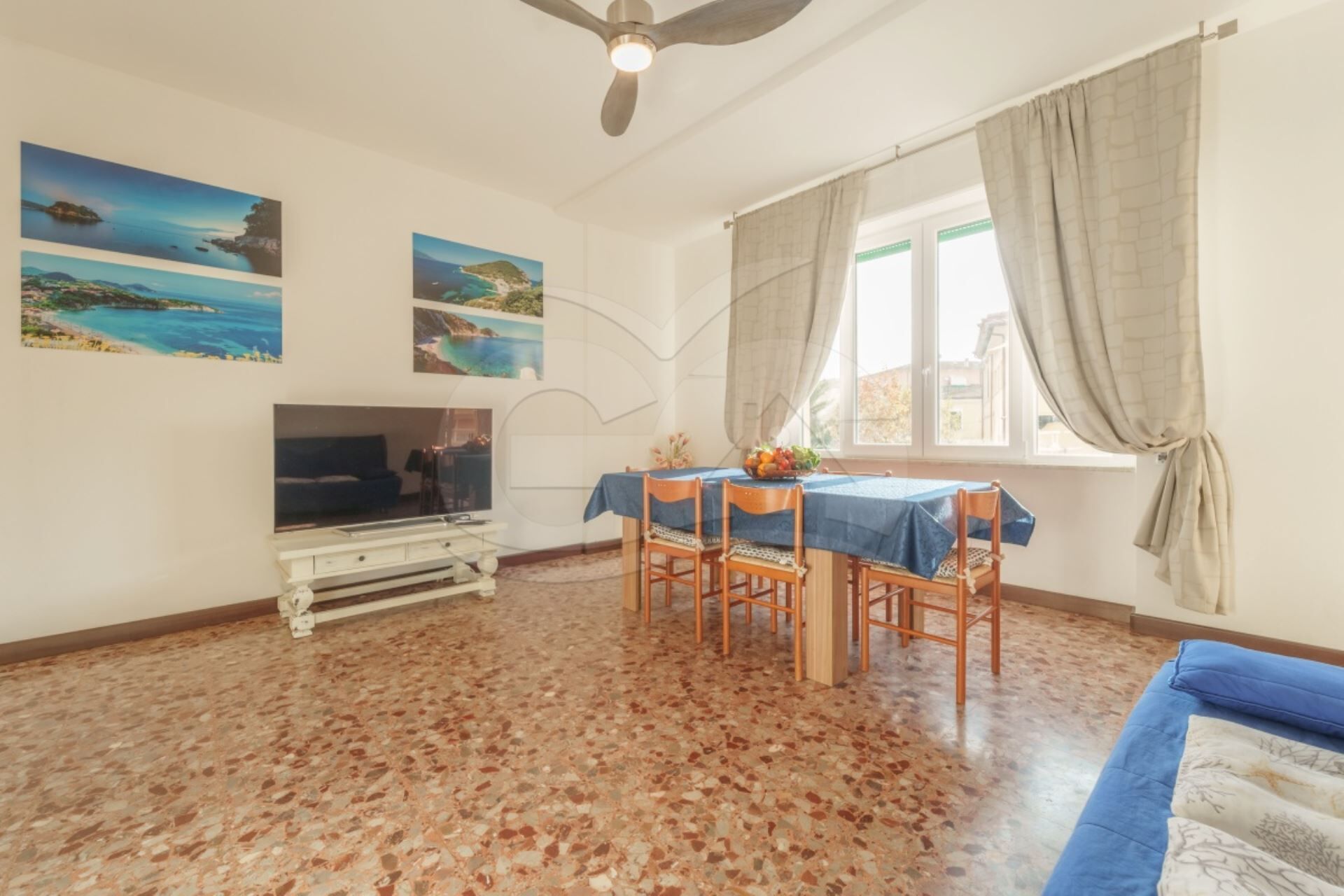 Un salon comprenant un téléviseur, une table à manger et des chaises, un ventilateur de plafond et une fenêtre avec des rideaux.