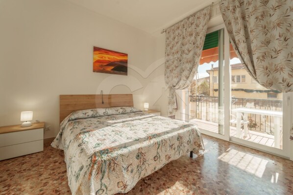 A bedroom with a bed, a nightstand, a painting, and a balcony with curtains.