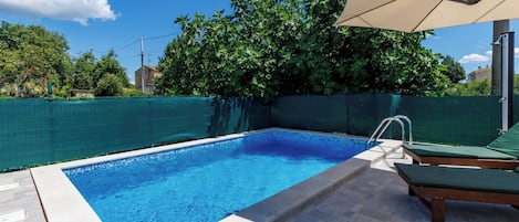 A swimming pool with a tiled deck, a lounge area with a canopy, and a green fence surrounding the area.