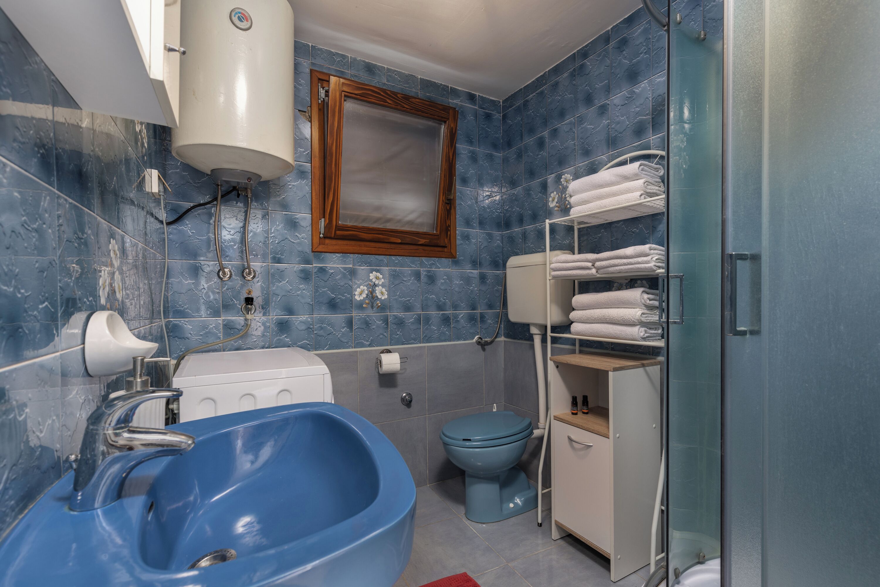 A bathroom with a blue sink, toilet, and shower. There are towels and toiletries on shelves.
