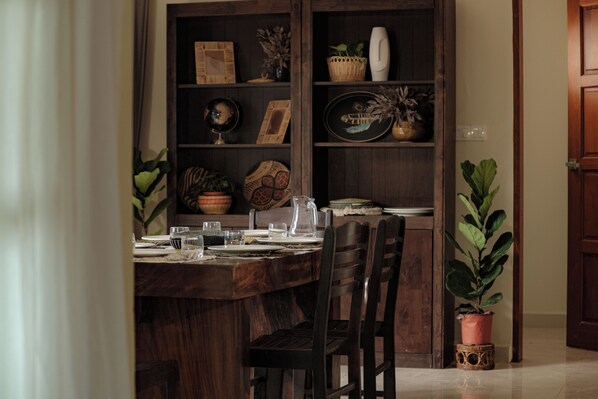 A dining area with a wooden table set for four, surrounded by dark wooden chairs. A wooden bookshelf with various decorative items is to the left. - Pickleball Hill Villa Langkawi (Langkawi)