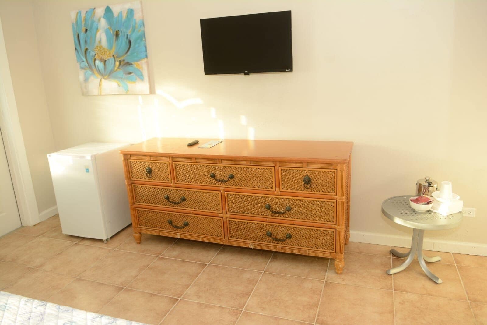 Una habitación con un aparador de madera con varios cajones, un refrigerador blanco pequeño, un televisor montado en la pared y una mesa redonda con dispensador de agua.