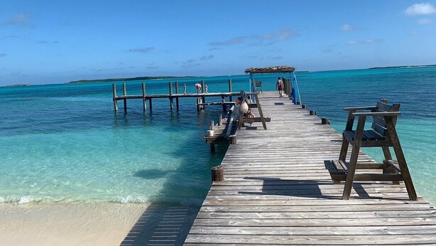 A wooden pier extending into clear turquoise waters with a few people and a thatched-roof structure at the end. - Exuma Beach Point (Rolleville)